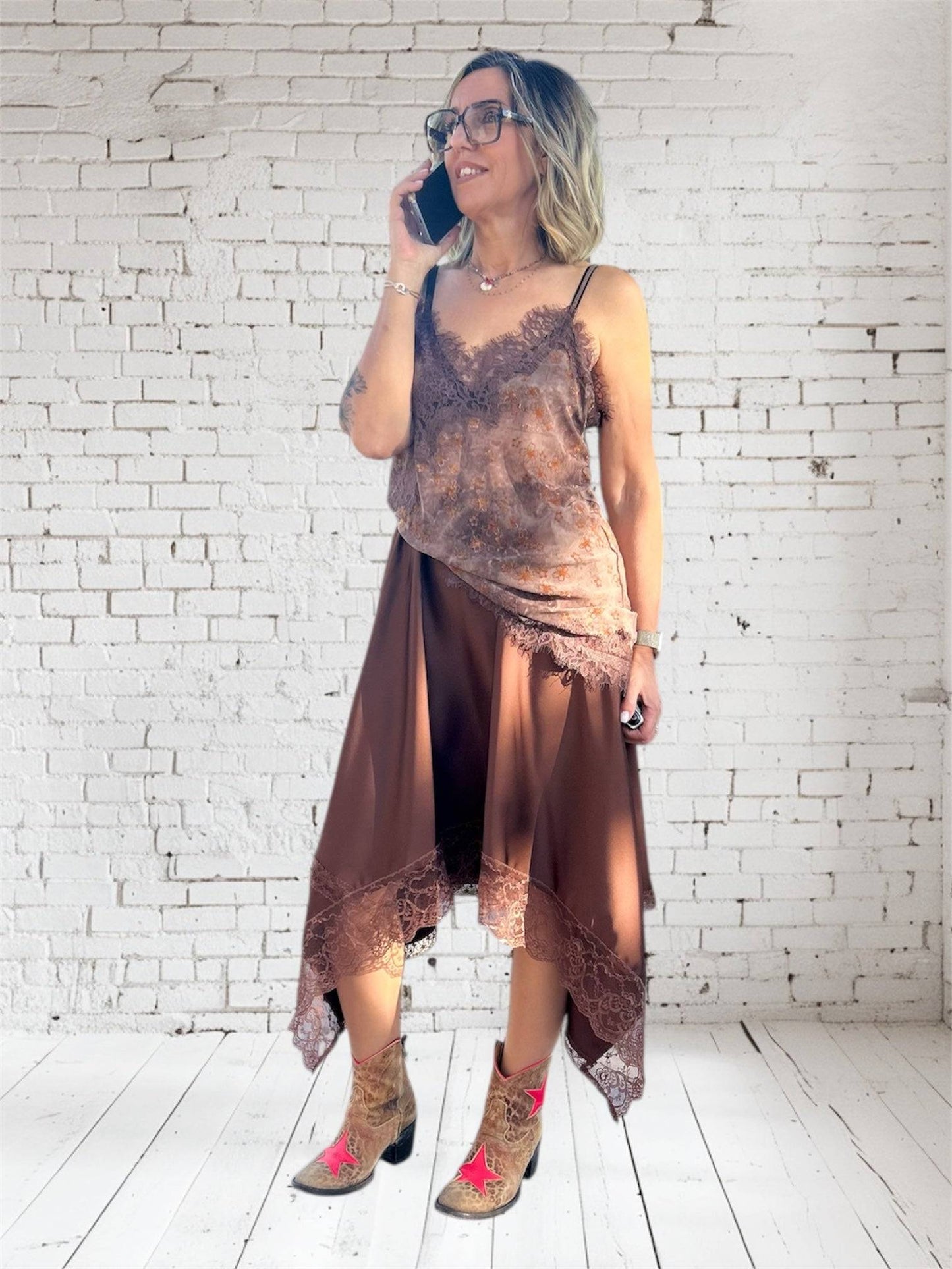 Jupe satinée Alya asymétrique chocolat avec pans longs et finition dentelle, portée avec un top assorti et des bottines western. Jupe tendance 2025 chic et fluide pour femme.
