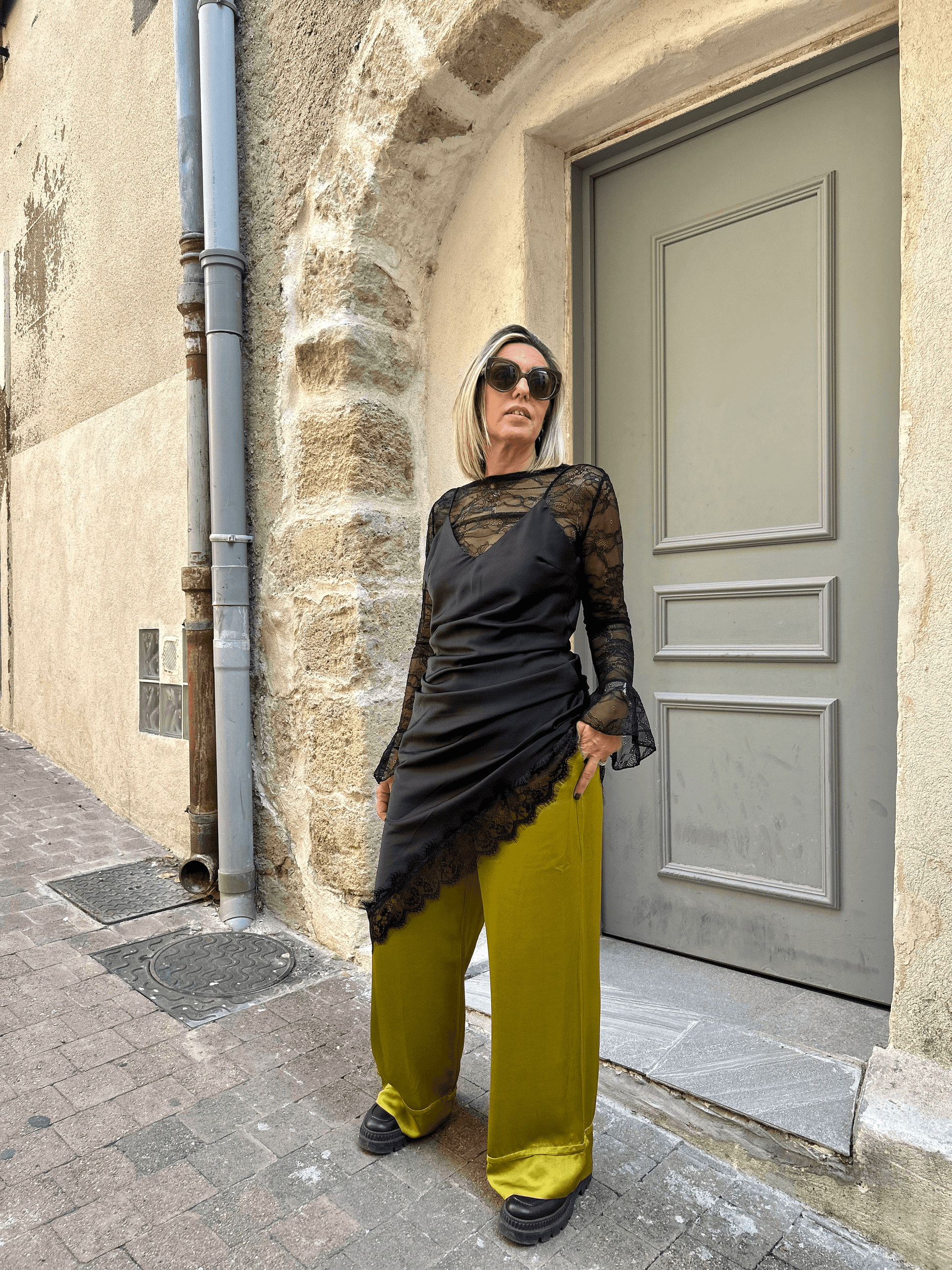 Robe noire asymétrique Carmen superposée à un top en dentelle noire et pantalon jaune moutarde, look urbain et audacieux pour femme élégante.