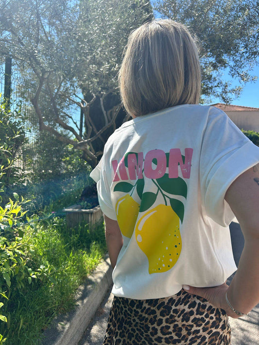 T-shirt Limoncello parfait pour l'été 2025, blanc avec imprimé citrons jaunes et lettrage rose, porté en extérieur sous un olivier.