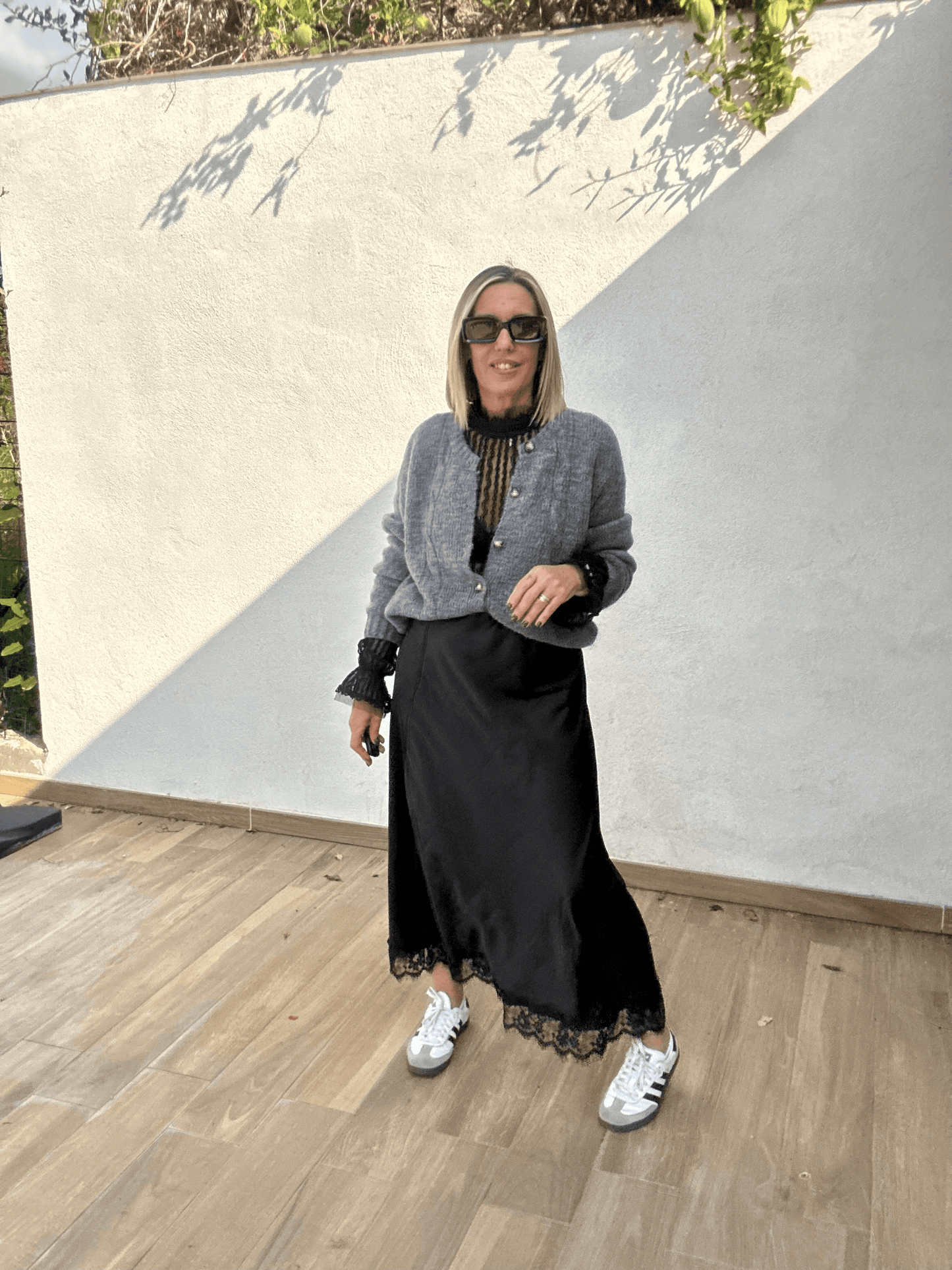 Jupe noire satinée femme Ziggy fluide avec finition dentelle, portée avec un cardigan gris et des baskets blanches, look chic décontracté automne-hiver 2025.