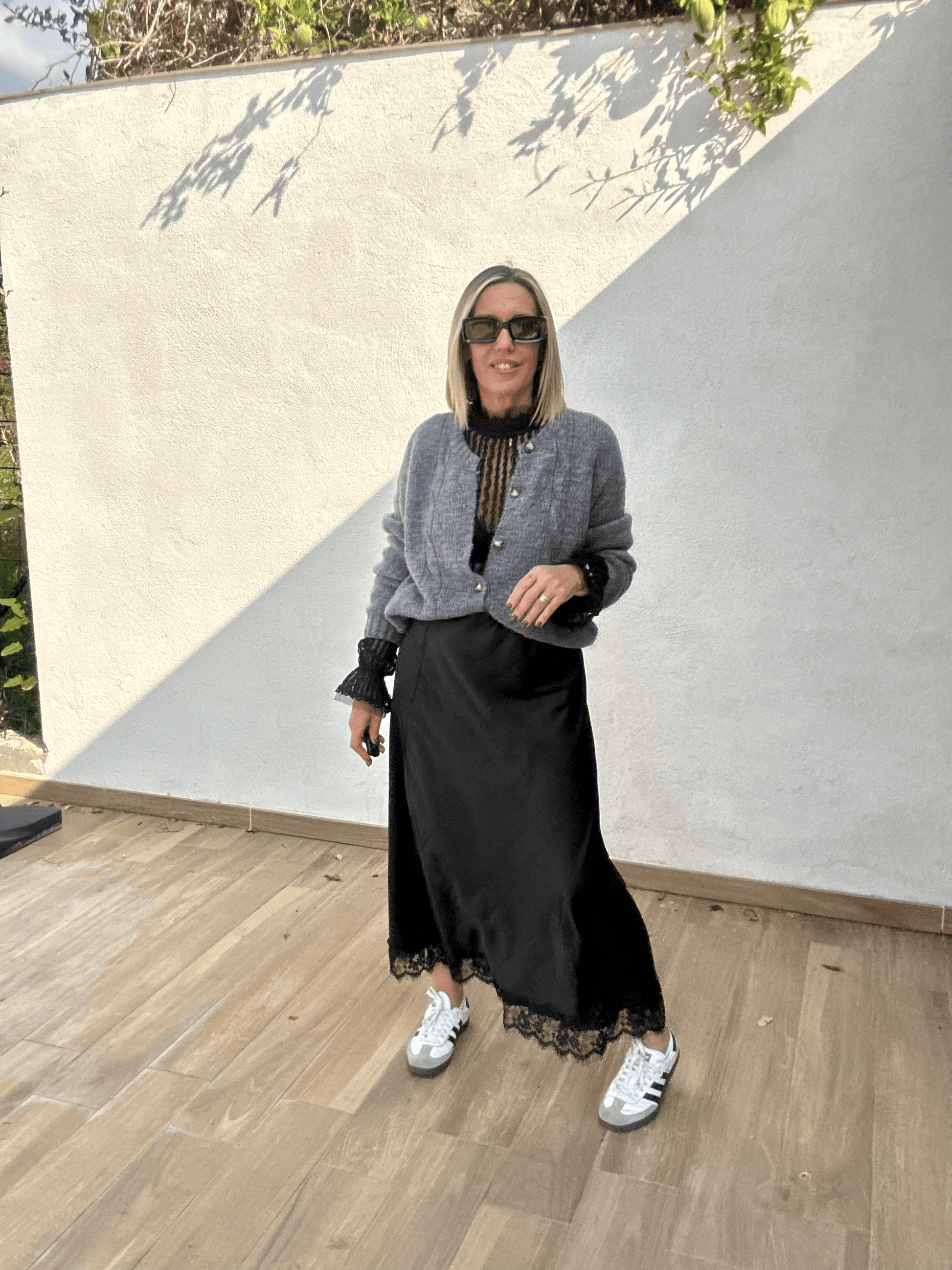 Jupe noire satinée femme Ziggy fluide avec finition dentelle, portée avec un cardigan gris et des baskets blanches, look chic décontracté automne-hiver 2025.