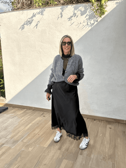 Jupe noire satinée femme Ziggy fluide avec finition dentelle, portée avec un cardigan gris et des baskets blanches, look chic décontracté automne-hiver 2025.
