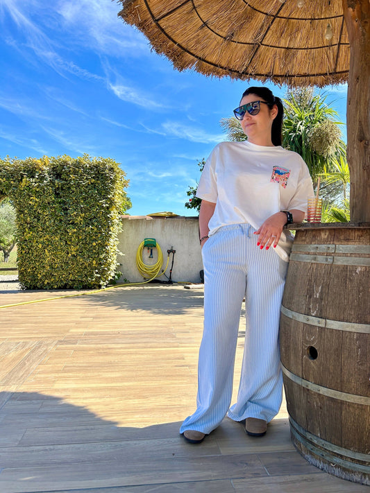 Pantalon Lido rayé bleu ciel et blanc porté avec un t-shirt blanc, idéal pour un look d'été 2025 chic et décontracté en bord de mer.